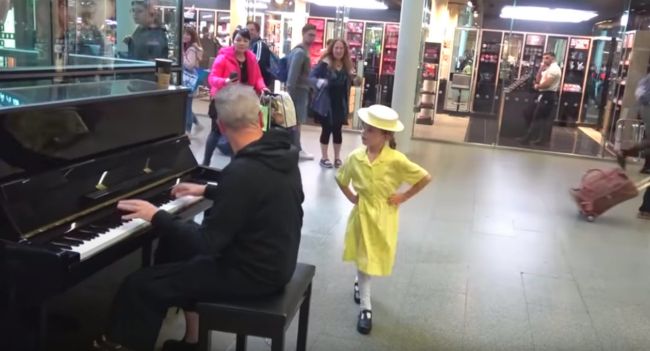 A little girl joins a man playing a piano in the street and starts dancing